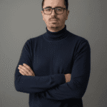 Marco Olmedi con sguardo serio, docevita blu scuro e braccia incrociate sul petto